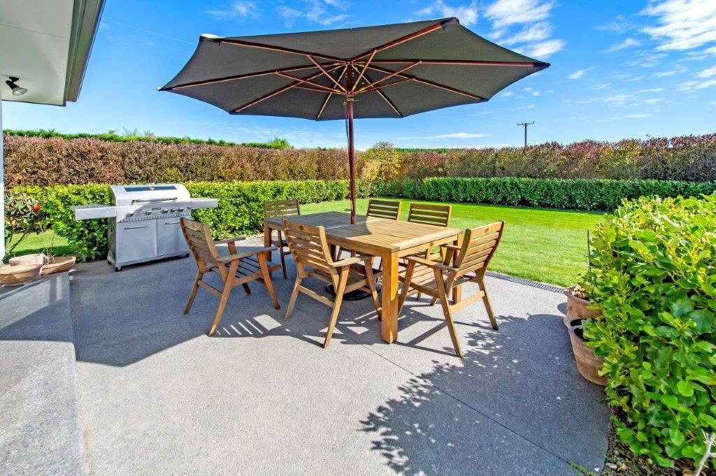 Paved outdoor dining area with a teak table, umbrella and barbecue, backed by photinia hedging
