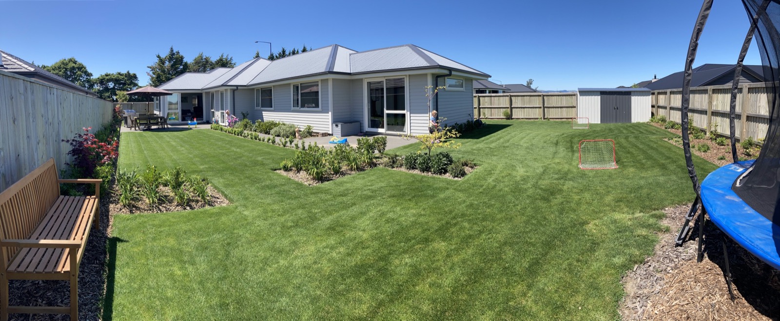 Wide view across the finished garden — lawn, planted beds, a bench and a trampoline